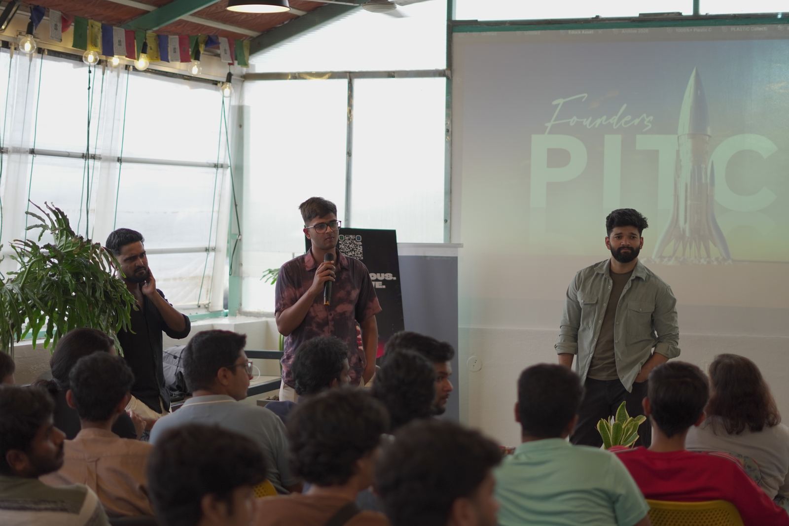 A founder pitching to a seated room, with the Founders Pitch title slide projected on the wall beside him.