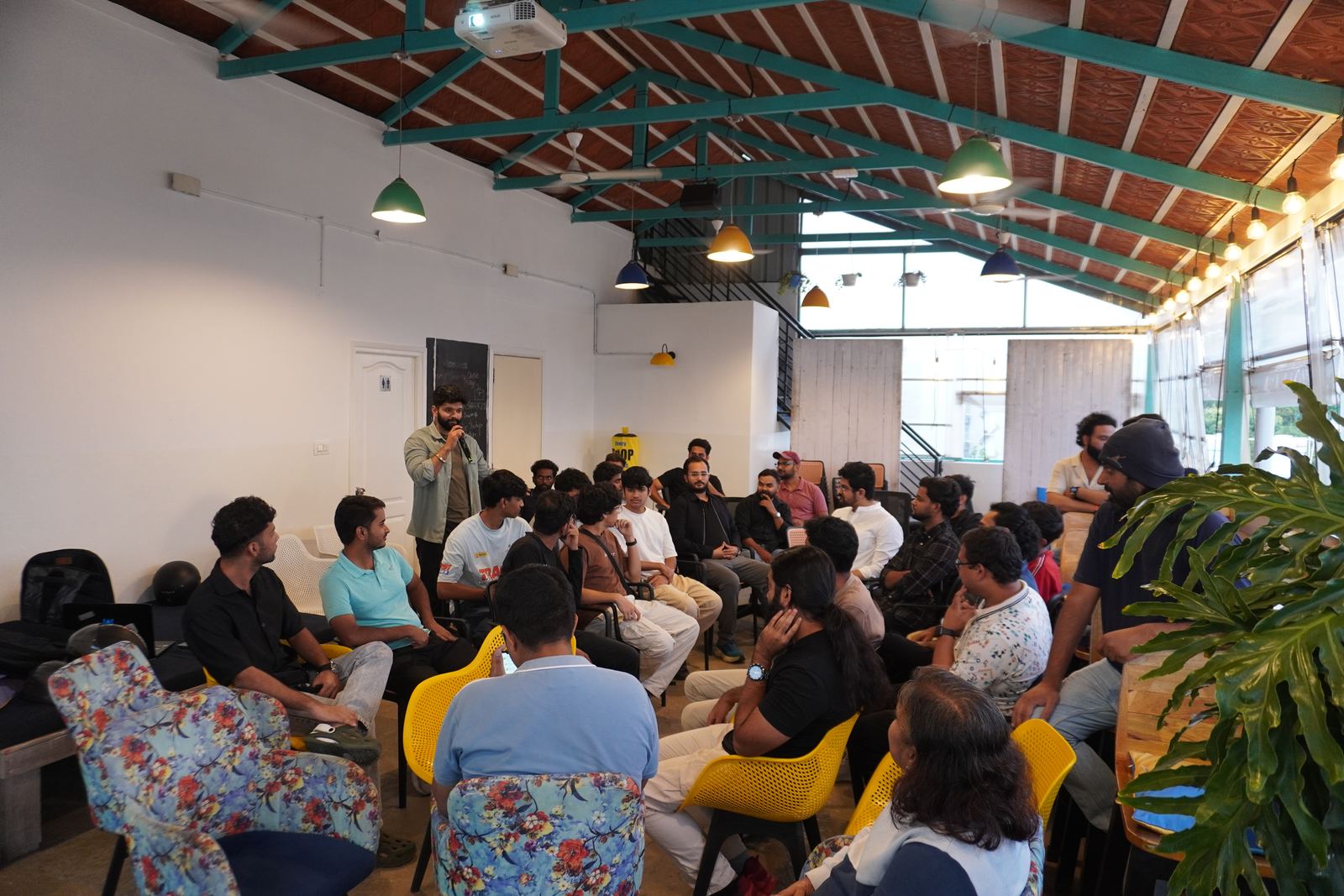 A full room at a past Edock meetup: founders seated in a circle of mismatched chairs under the venue's exposed teal roof trusses, listening to someone speaking with a microphone.