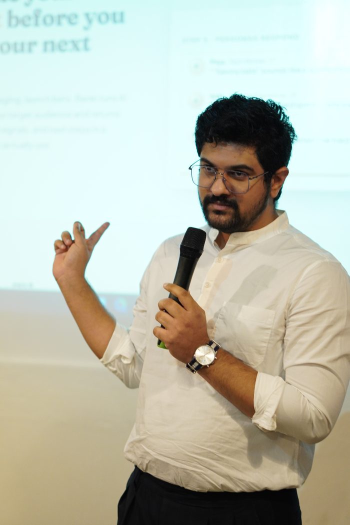 A founder mid-sentence with a microphone, gesturing to a slide projected on the wall behind him.