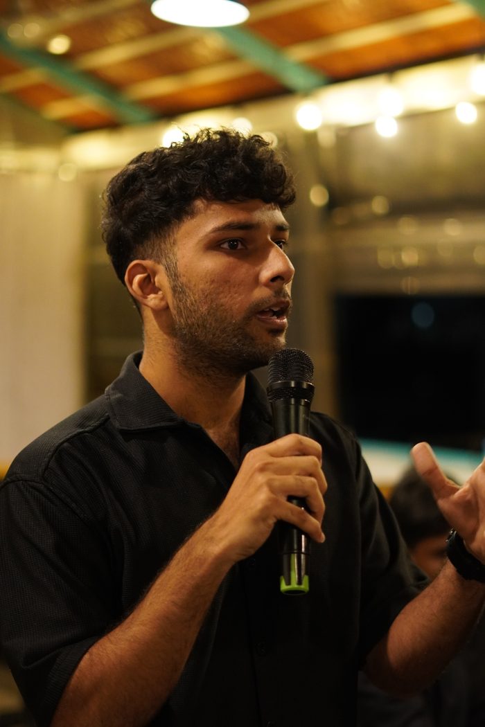 A founder speaking into a handheld microphone in the evening, string lights strung across the roof behind him.