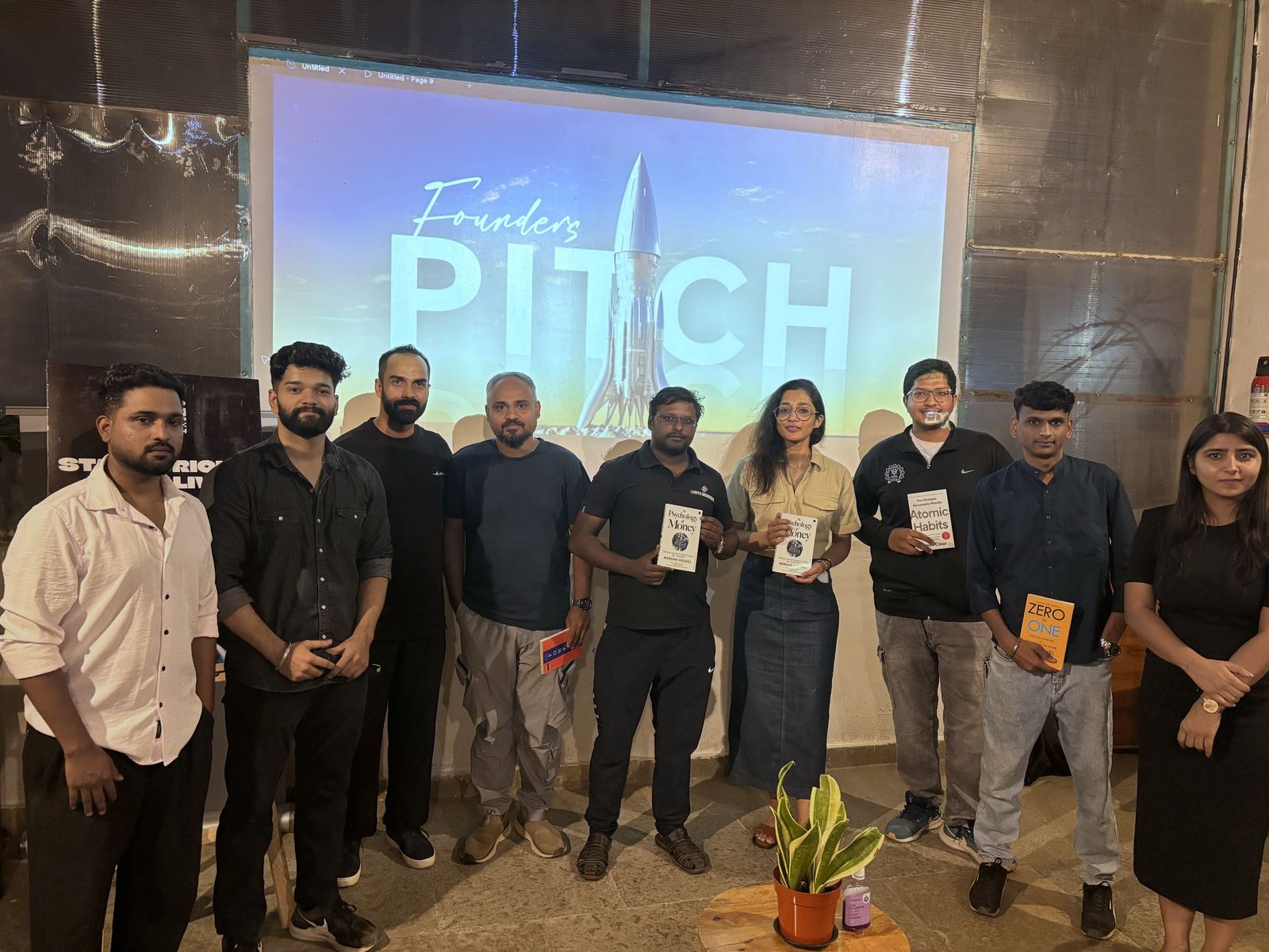 A group at a past Founders
            Pitch standing in front of the projected title slide, several holding books.